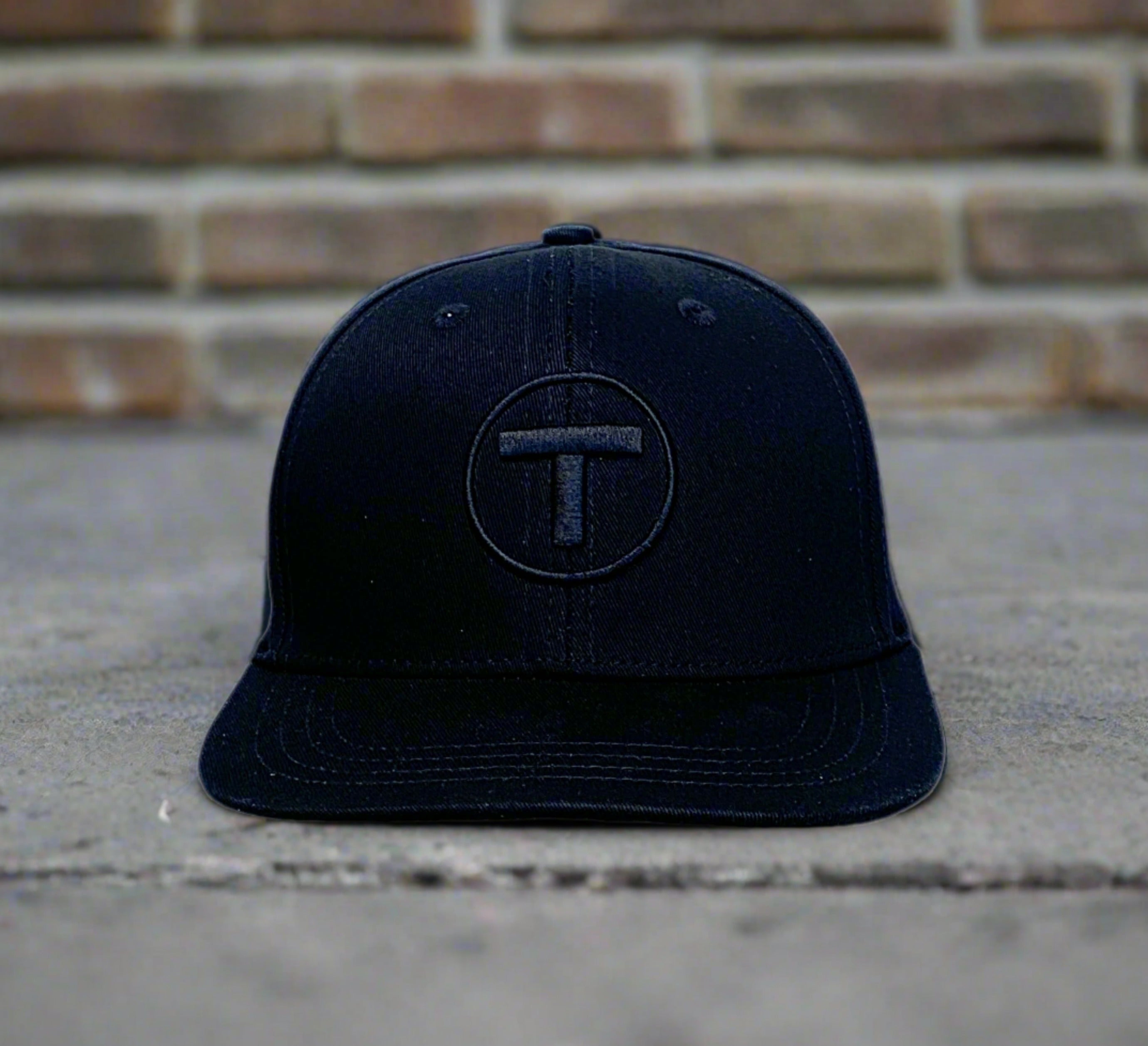 Front view of black flat brim youth cap with puff embroidered MBTA T Logo